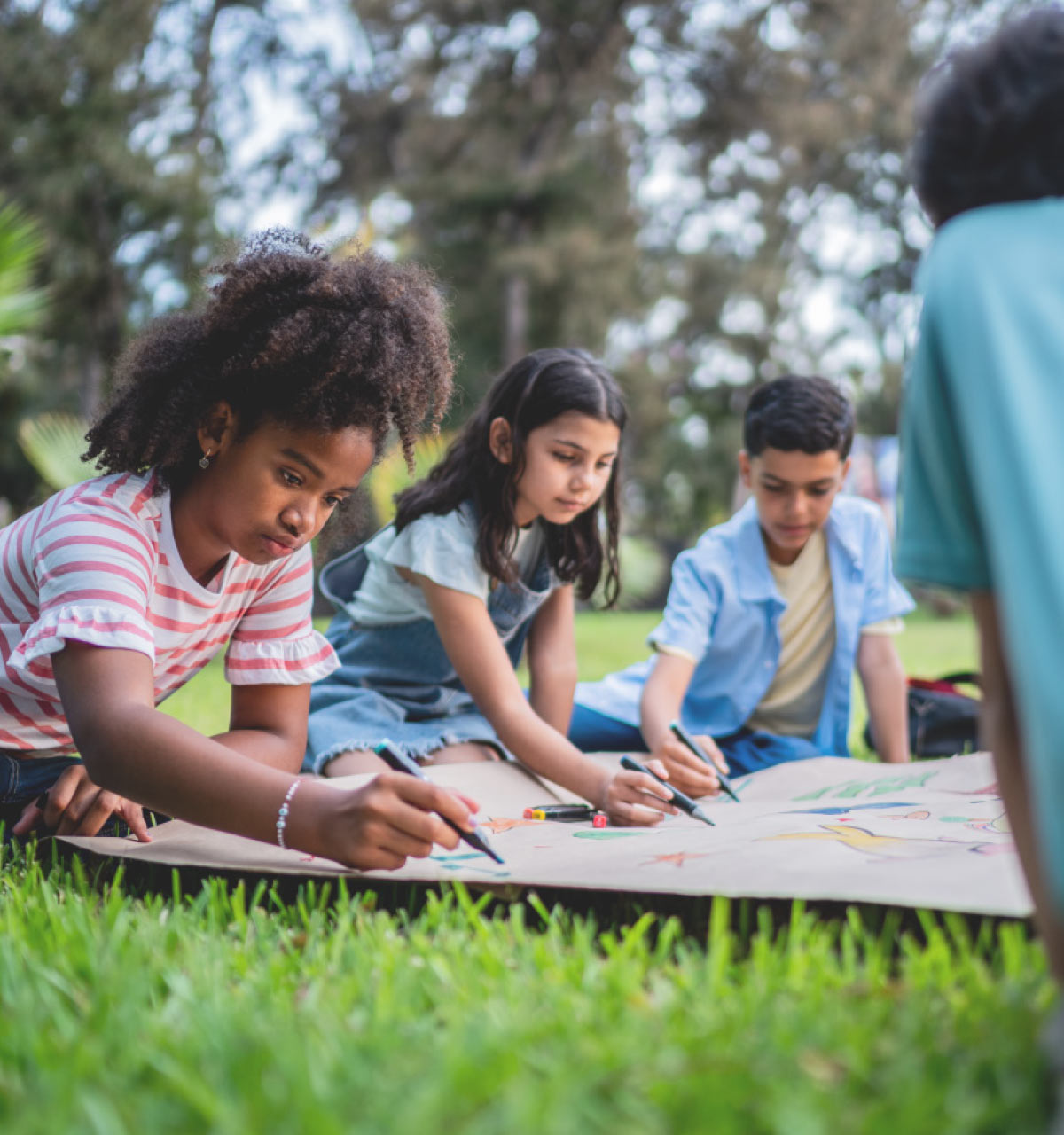 Diverse group of children sitting on grass, focused on drawing together on a large sheet of paper with markers in an outdoor setting.