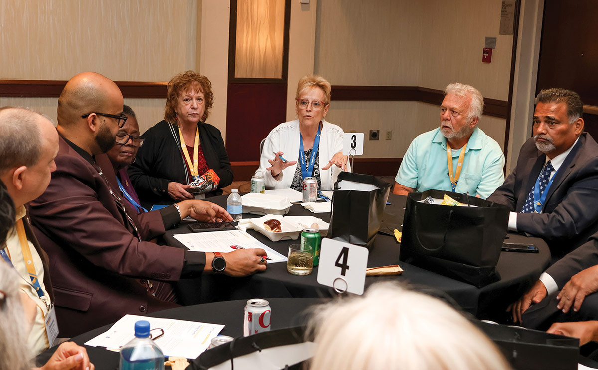 A group of conference attendees sit around a round table engaged in discussion. A woman in a white blazer gestures while speaking as others listen. Papers, soda cans, and table numbers are visible on the black tablecloth.