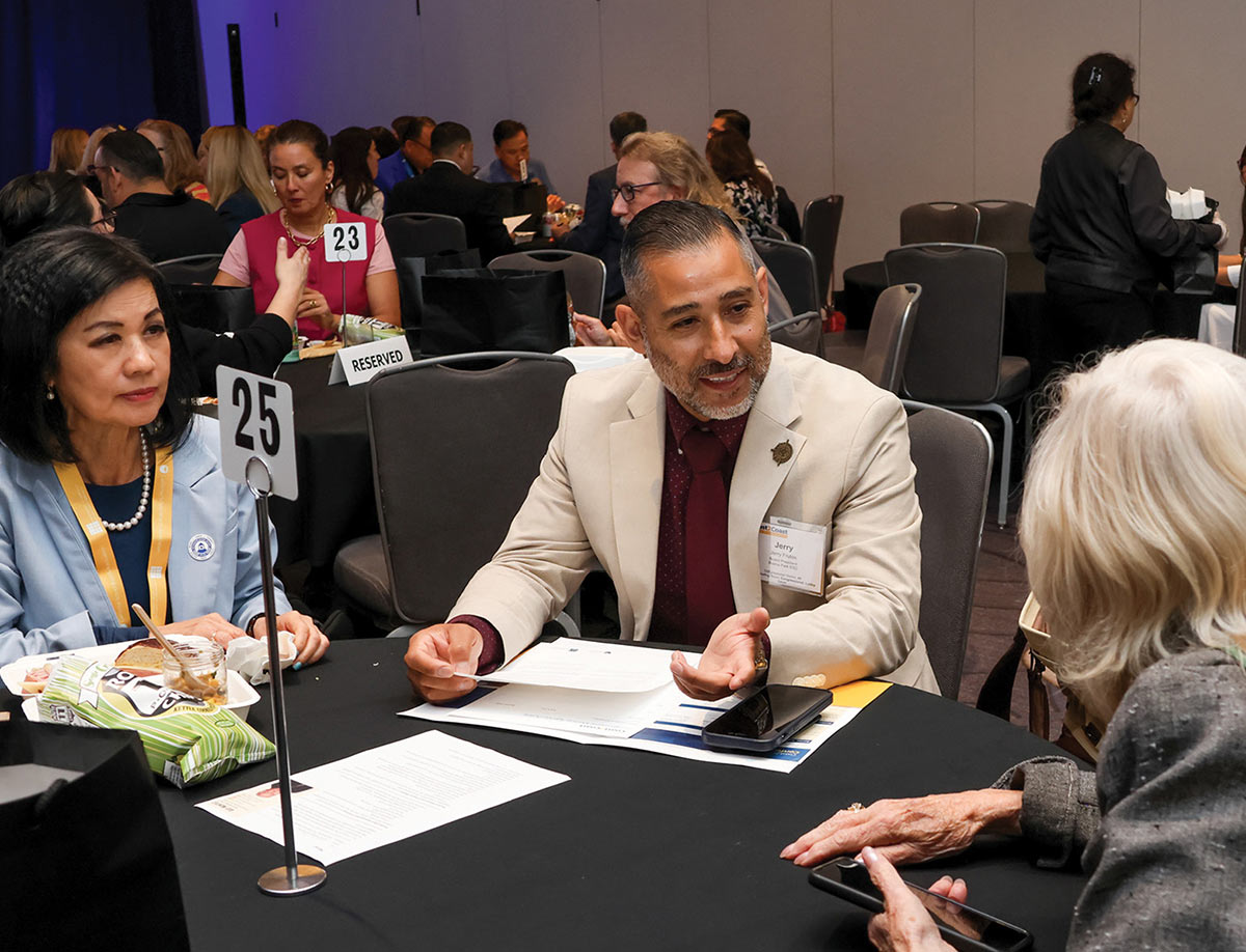 A man in a beige suit and maroon tie speaks with other attendees at a round conference table marked with table number 25. A woman in a light blue blazer sits next to him, and papers are spread across the black tablecloth.