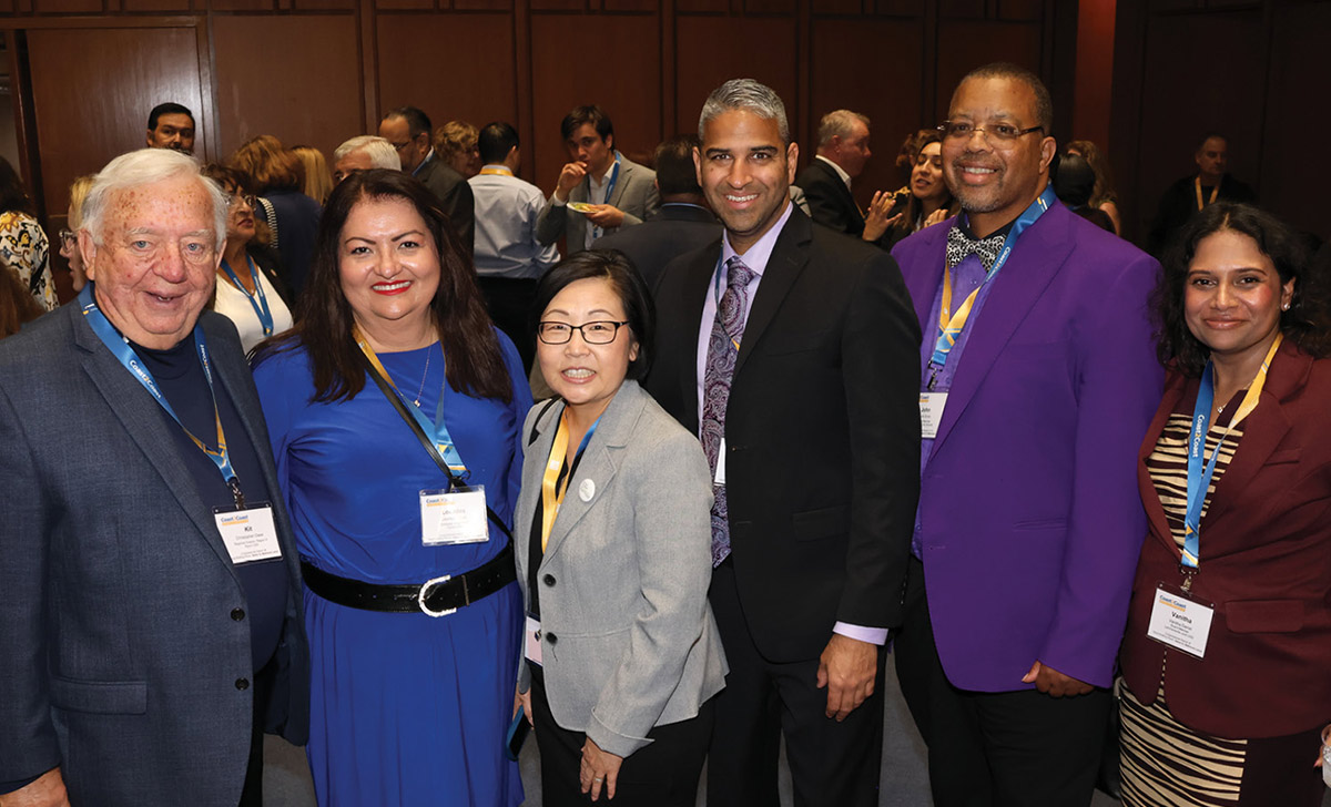 Six diverse conference attendees stand together and smile for a photo. They wear professional business attire—including a royal blue dress, a gray blazer, and a bright purple jacket—along with matching event lanyards.