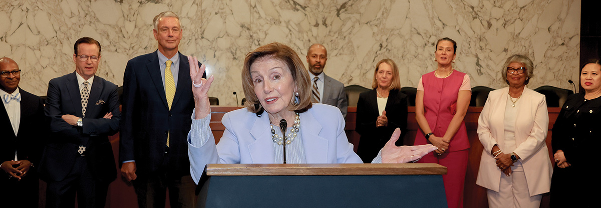 Nancy Pelosi speaks and gestures at a wooden podium. A group of professionally dressed men and women stand behind her against a light marble wall.