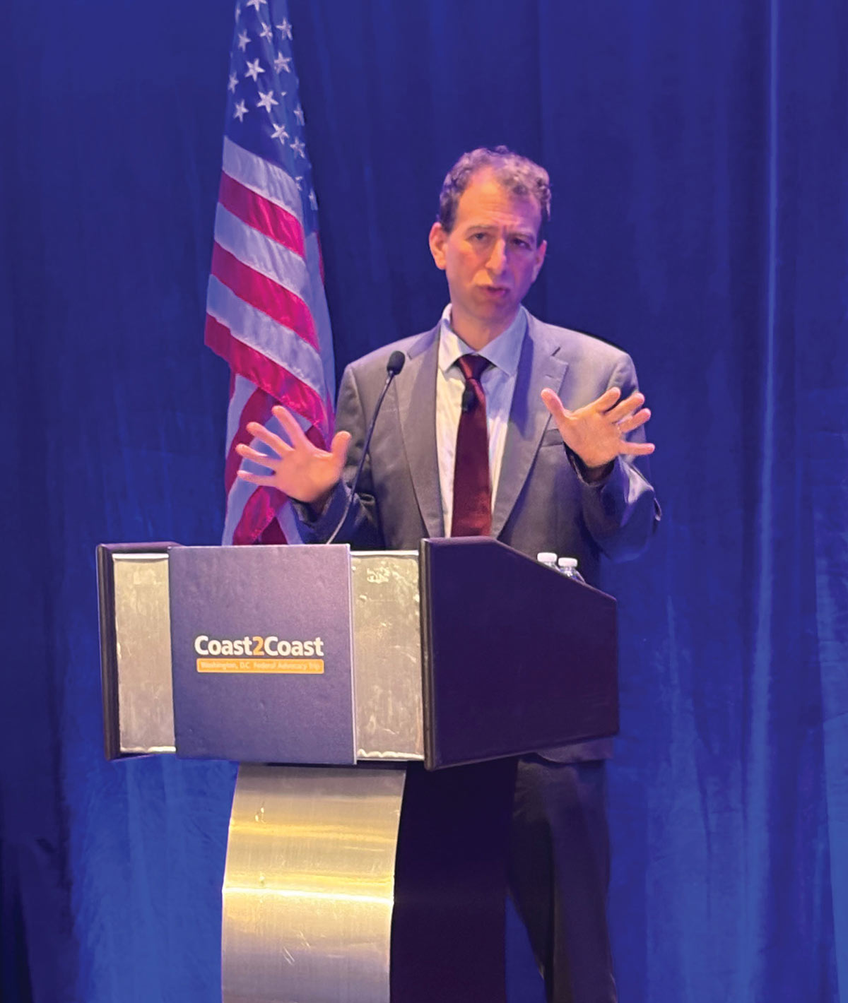A man in a gray suit and maroon tie gestures with both hands while speaking at a podium labeled "Coast2Coast." An American flag stands behind him against a deep blue curtain.