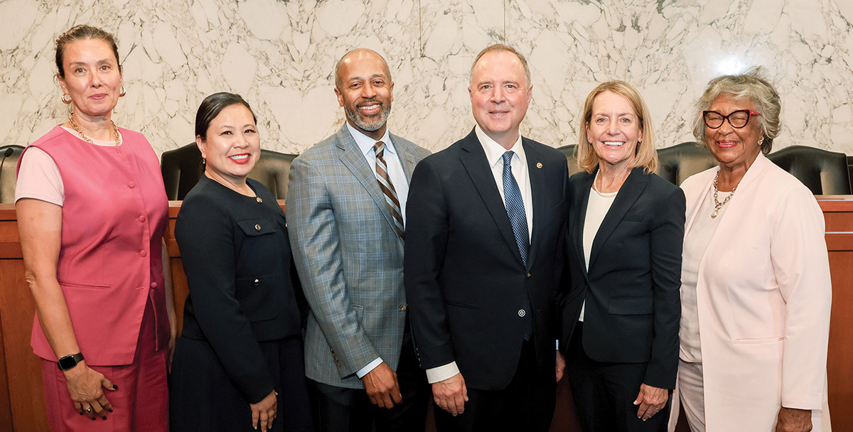 Six professionally dressed men and women stand in a line and smile for a photo. They wear business attire, including a fuchsia top, dark suits, and a plaid blazer, in front of a light marble wall.