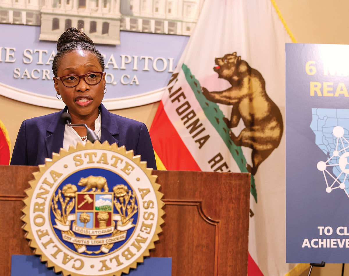 A woman wearing glasses and a navy blazer speaks at a wooden podium bearing the California State Assembly seal. The California state flag and a State Capitol sign are visible in the background.