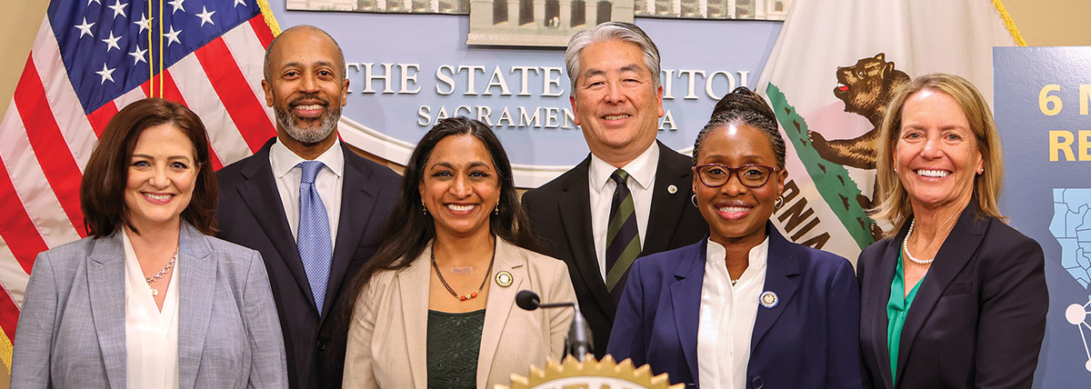 Six professionally dressed men and women smile for a group photo behind a press podium. The American flag, California state flag, and a "The State Capitol" sign are visible in the background.