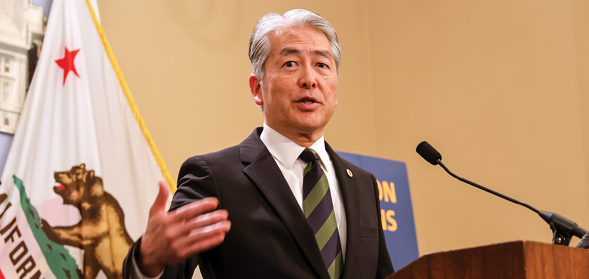 A man with graying hair in a dark suit and a striped tie speaks and gestures at a wooden podium with a microphone. The California state flag stands in the background.
