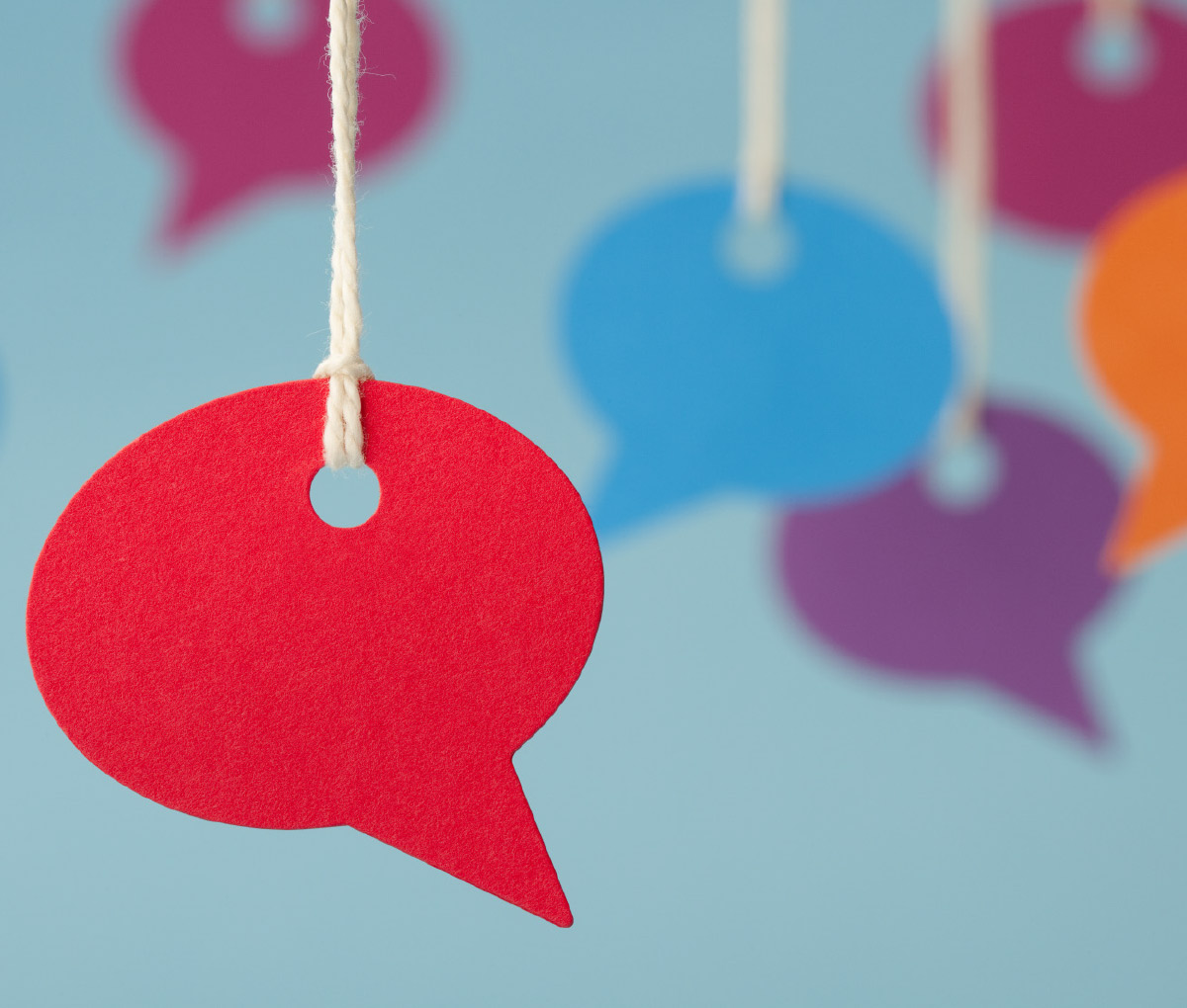 A close-up of a red paper speech bubble hanging by a string, with several colorful, blurred speech bubbles in the background against a blue surface.