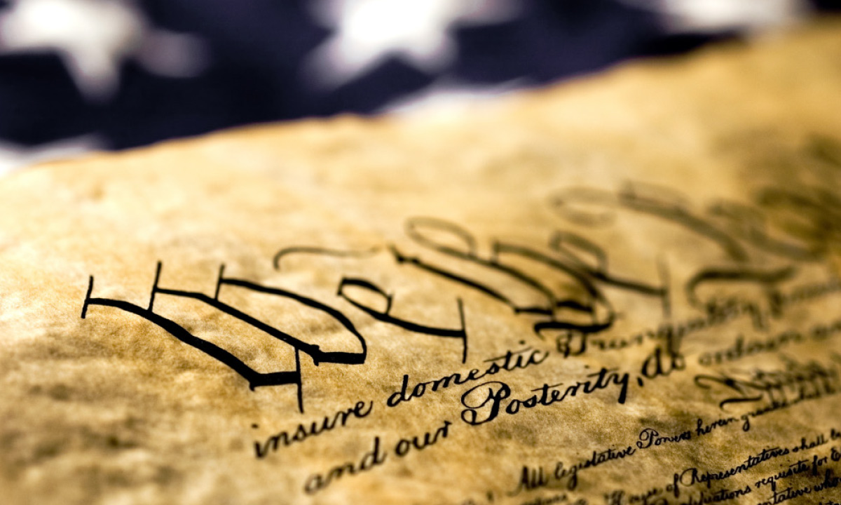A close-up, angled shot of the U.S. Constitution's preamble, "We the People," on aged parchment with an American flag blurred in the background.