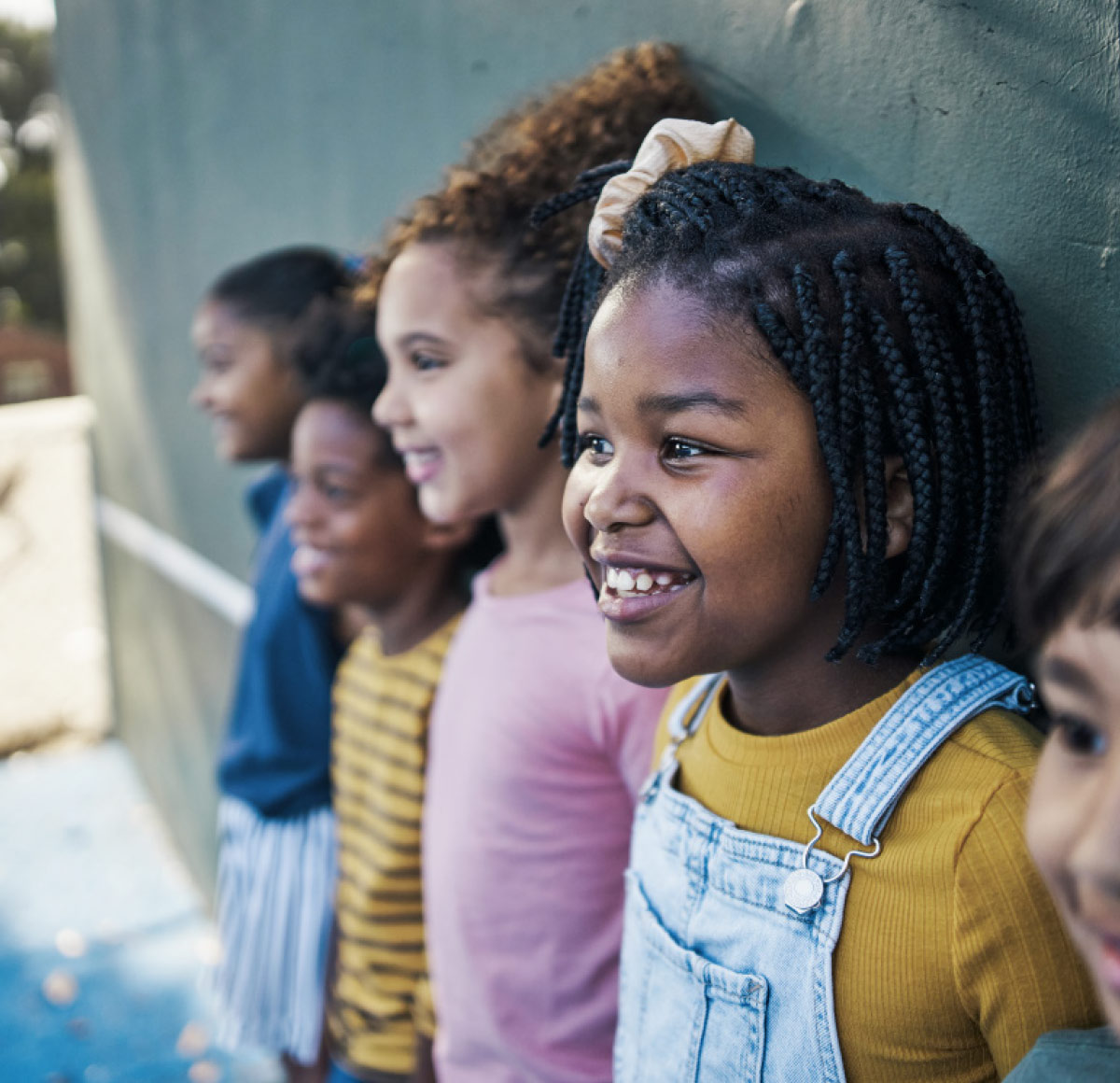 A diverse group of smiling children stand in a line against a green wall, with a young girl in denim overalls in the foreground.