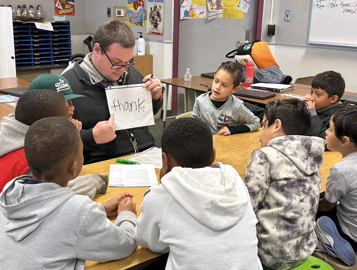 A teacher sitting at a classroom table with several young students, pointing to the handwritten word "thank" on a handheld sheet.