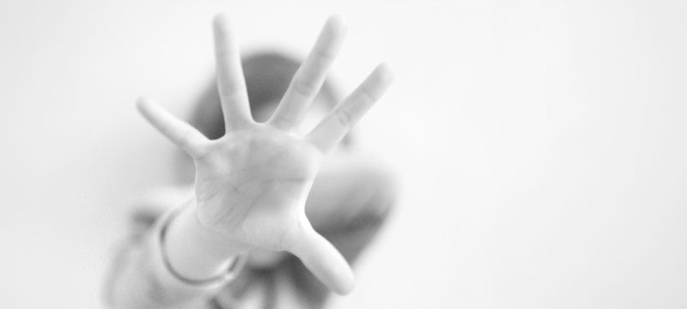 A blurred black and white photograph of a person holding their palm out toward the camera with fingers spread in a “stop” gesture.