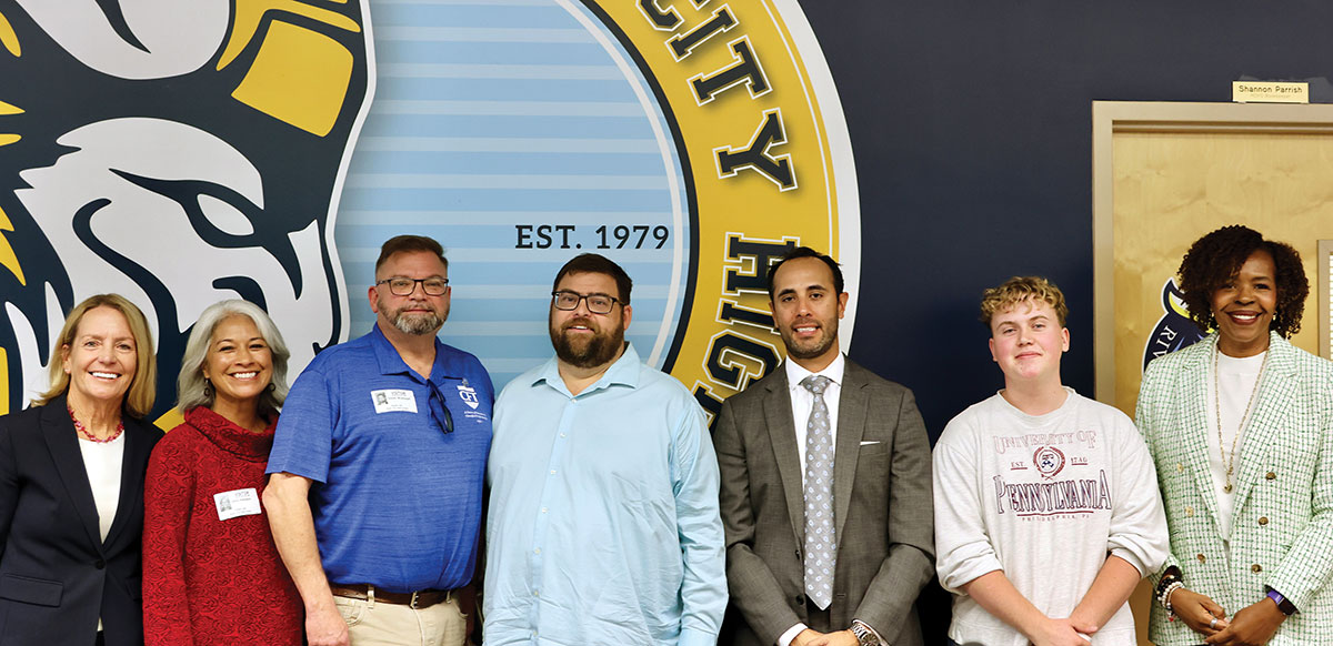 Group of educators and students posing in front of school emblem.