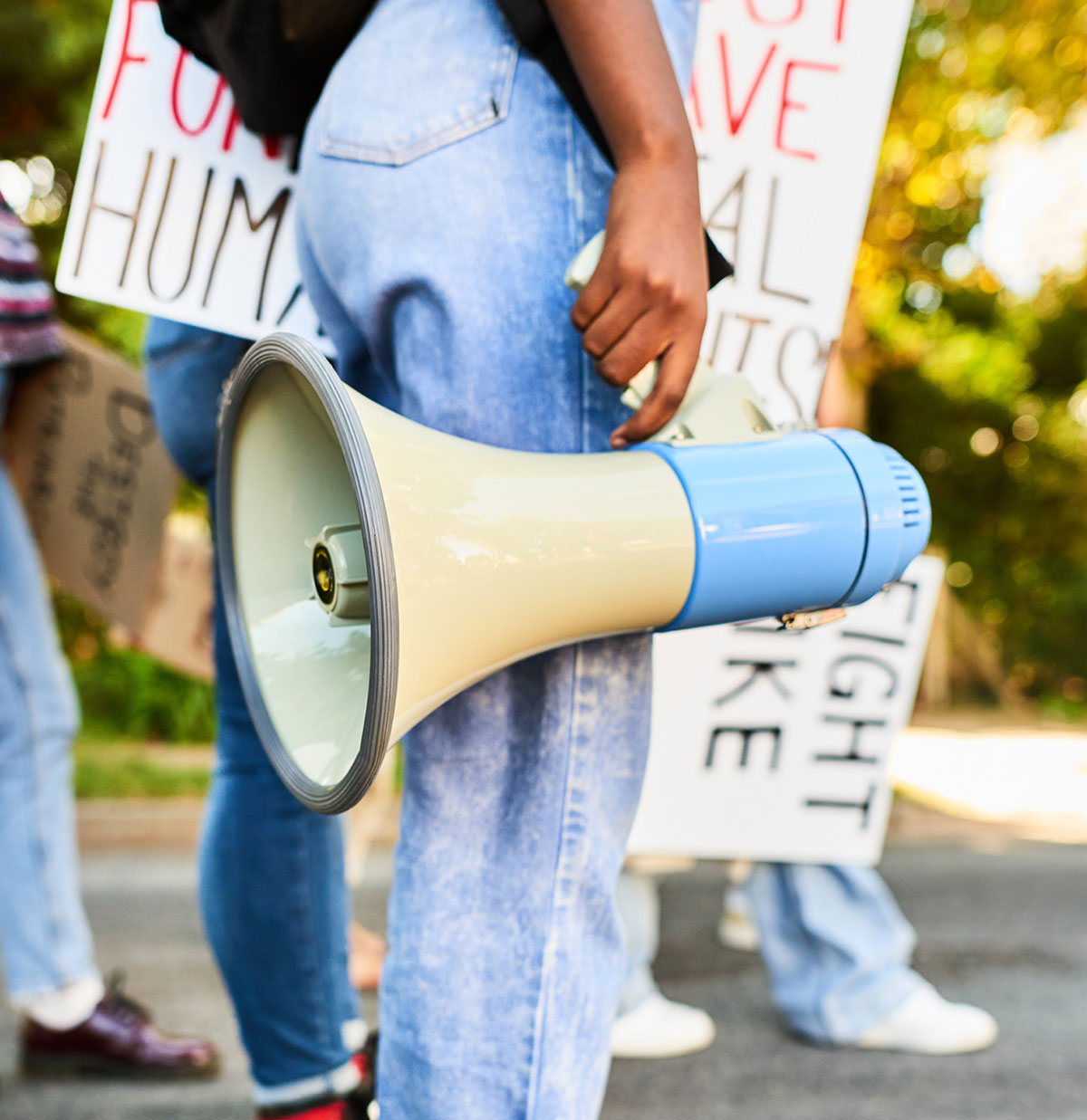 A close-up, low-angle shot of a demonstrator carrying a megaphone at an outdoor protest with other people nearby holding different protest signs