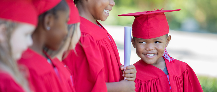 A smiling young boy in a red graduation gown and cap looks at his classmates.
