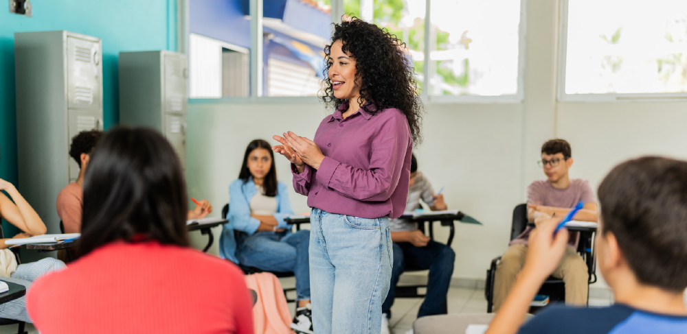 A smiling teacher with curly hair stands at the front of a classroom speaking to a diverse group of students seated at their desks.