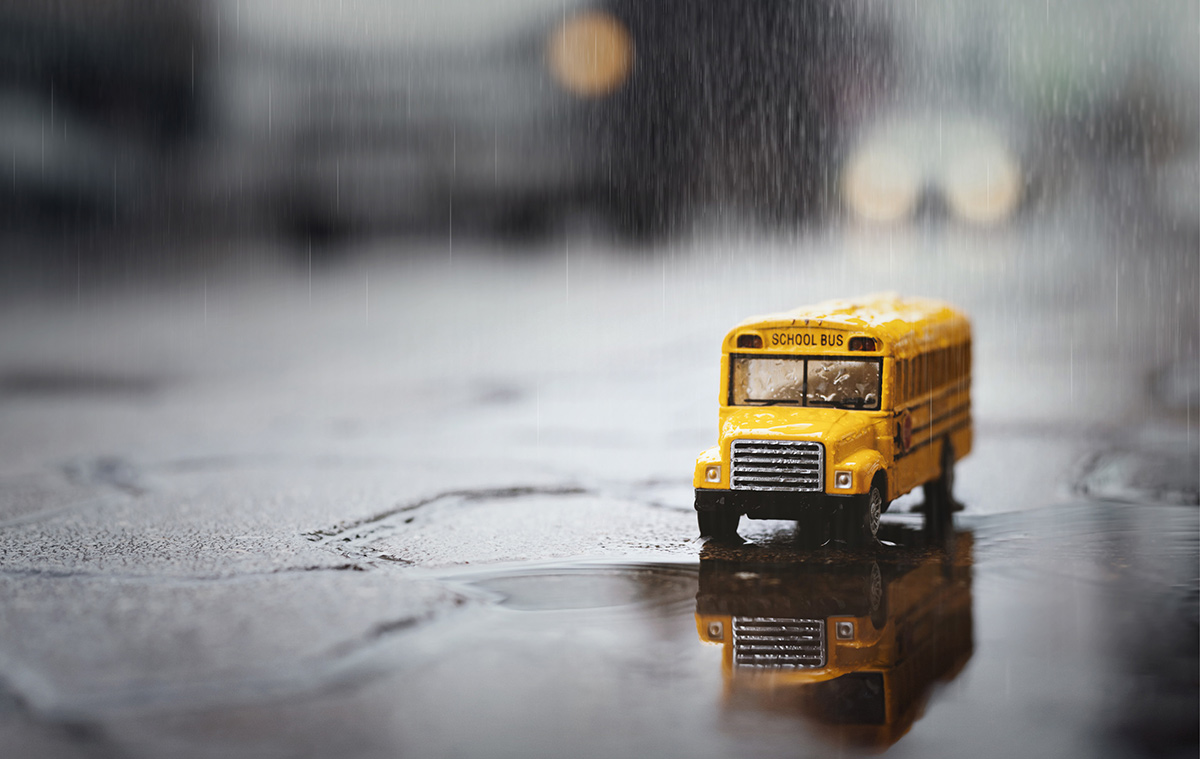 Small, yellow toy school bus on a wet, rainy pavement, creating a clear reflection in a shallow puddle while streaking rain pours