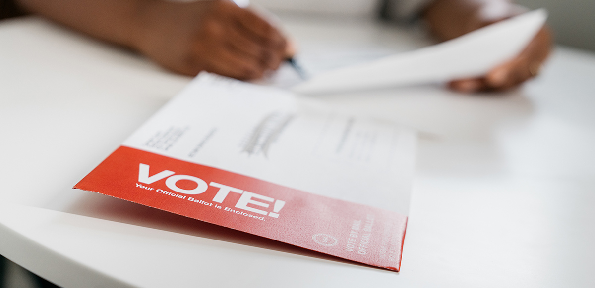 A person fills out a mail-in ballot behind an envelope marked "VOTE!".
