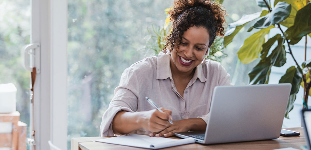 A smiling woman with curly hair taking notes on a notepad next to a open laptop.