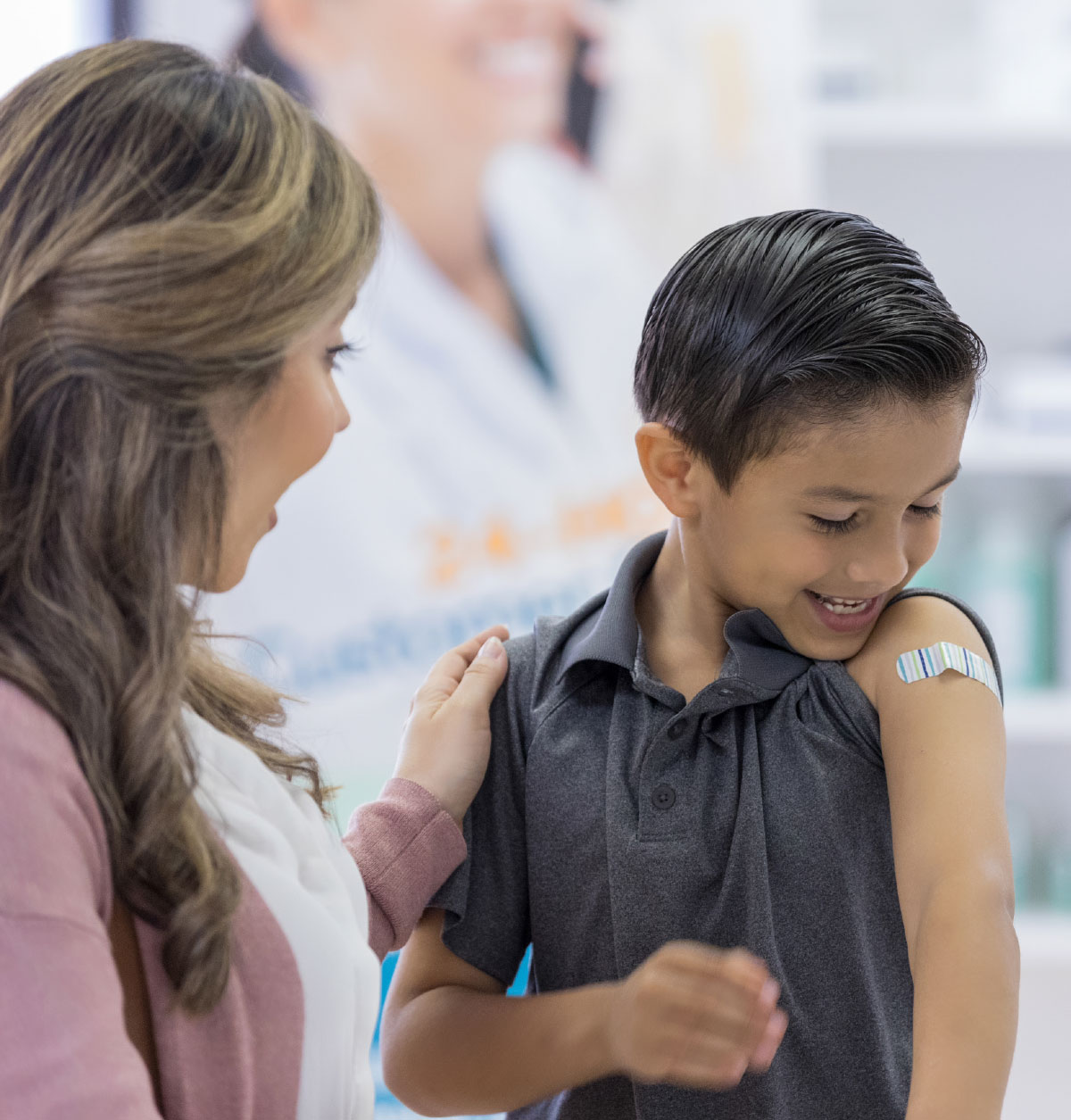 A smiling boy looks down at a striped bandage on his upper arm while a mother comforting him rests a hand on his shoulder.