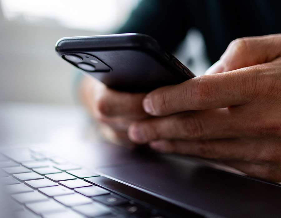 A person holding and typing on a black smartphone above a laptop keyboard.
