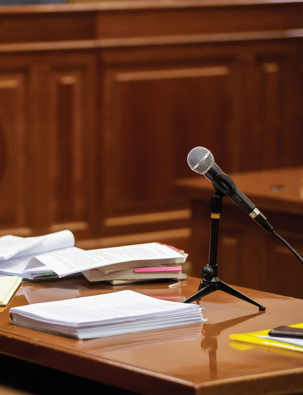 A microphone on a small tripod stand next to stacks of documents on a wooden courtroom table.