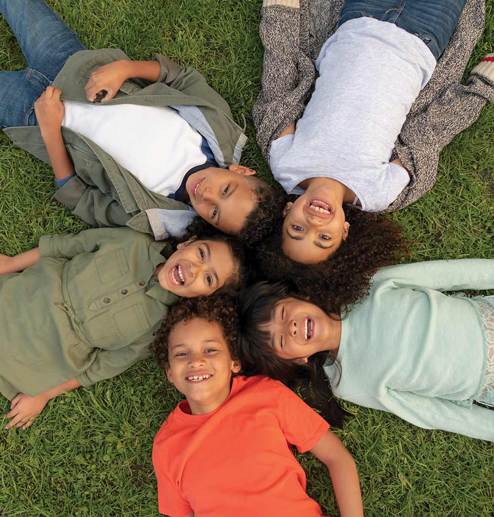 Five children lying in a circle on green grass, smiling up at the camera.
