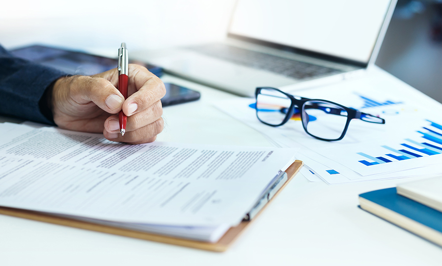 A person holds a red pen writing on paperwork at a desk next to glasses and charts.