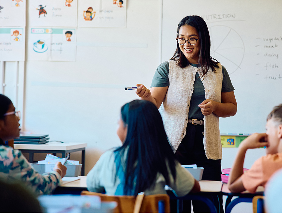 A teacher wearing glasses pointing with a marker while addressing students in a classroom.