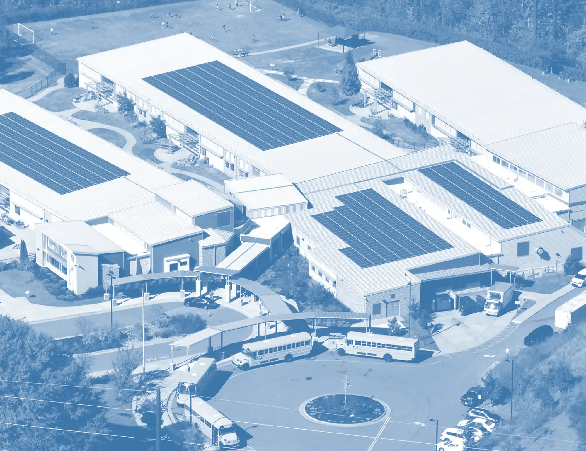 Blue-toned aerial view of a school campus with rooftop solar panels, walkways, and yellow school buses lined up.