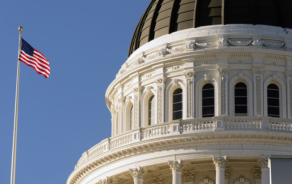 Close-up of an ornate white capitol building dome featuring arched windows and columns, with an American flag flying against a clear blue sky.