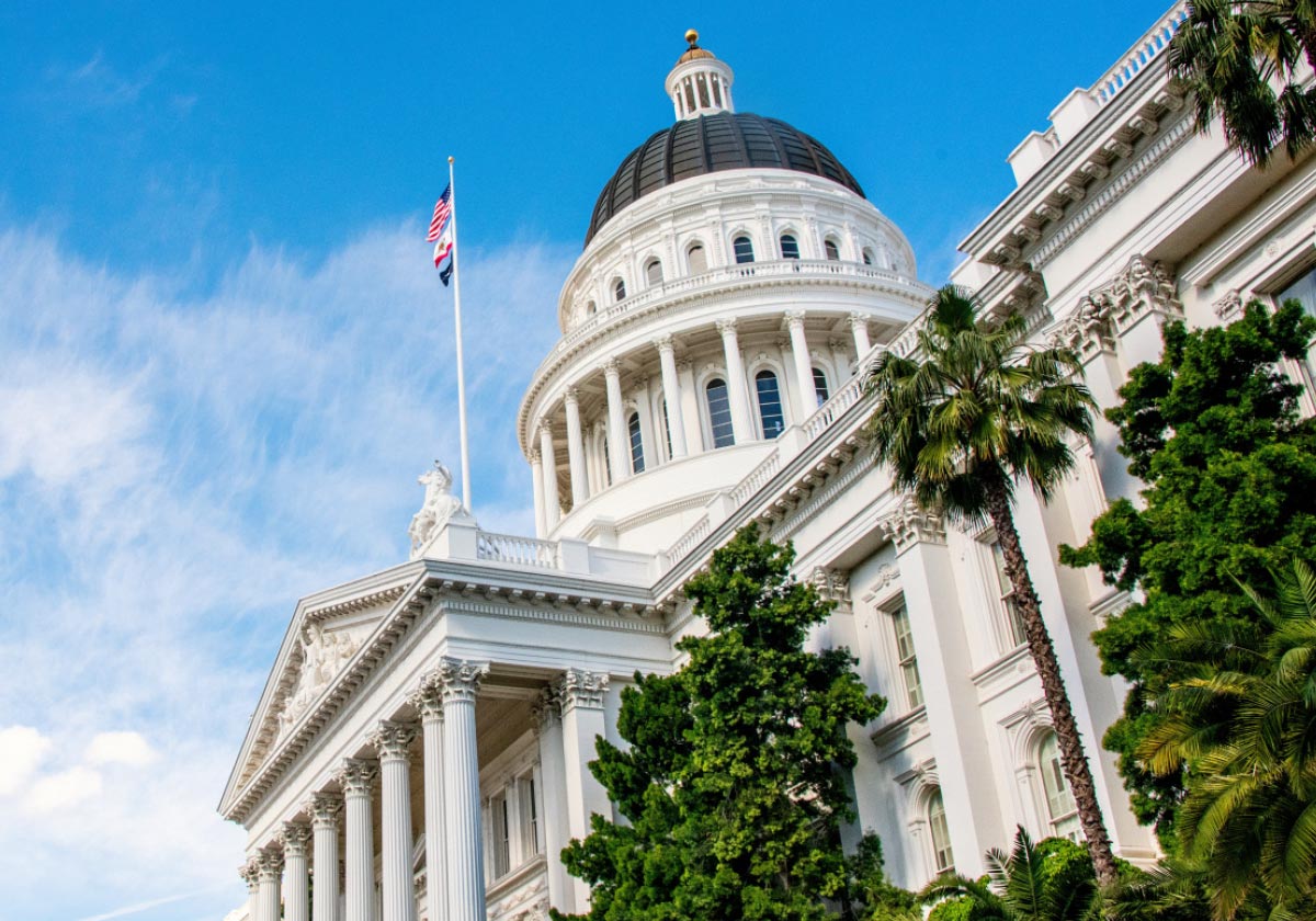 Low-angle view of the California State Capitol dome and white columns framed by palm trees against a blue sky.