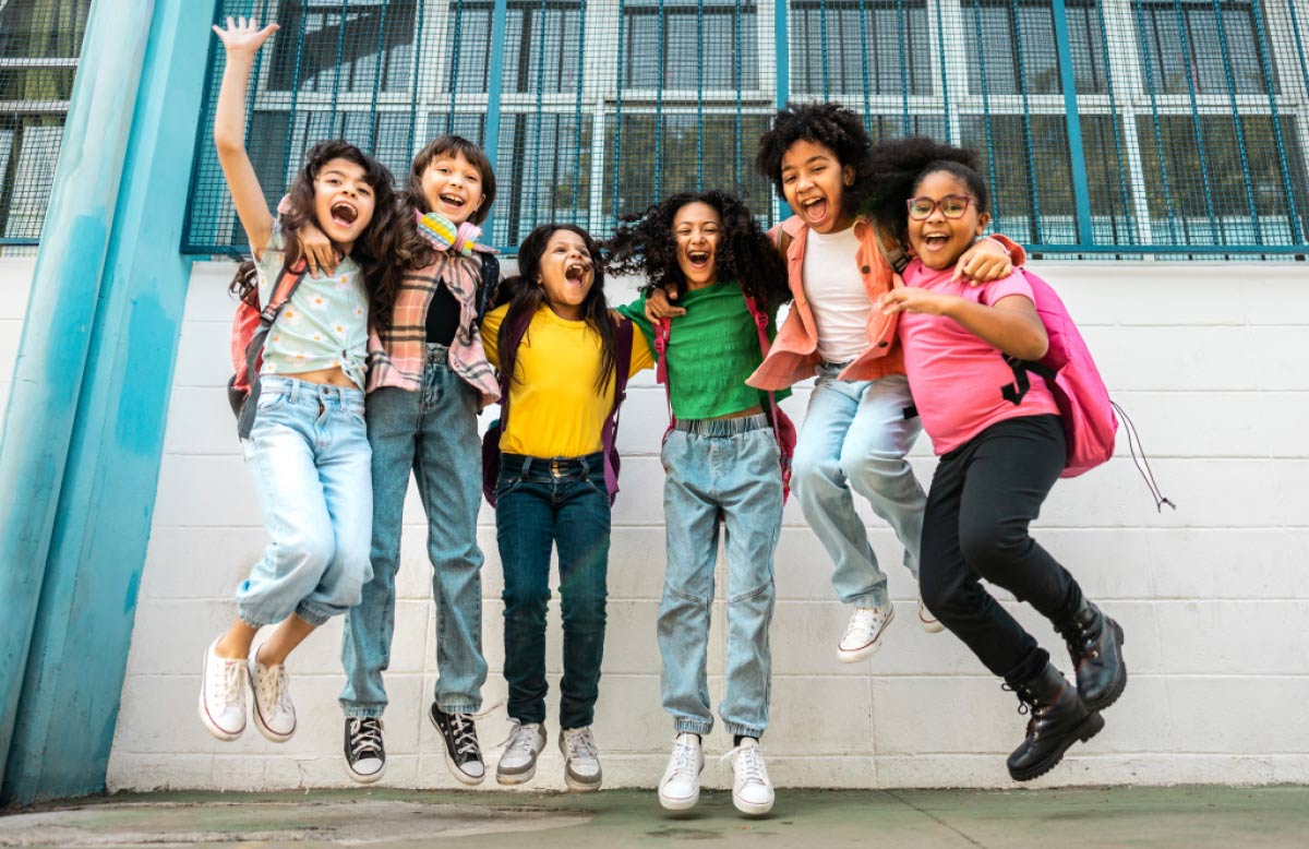 Group of six joyful young students in colorful outfits jumping in the air in front of a school building wall.
