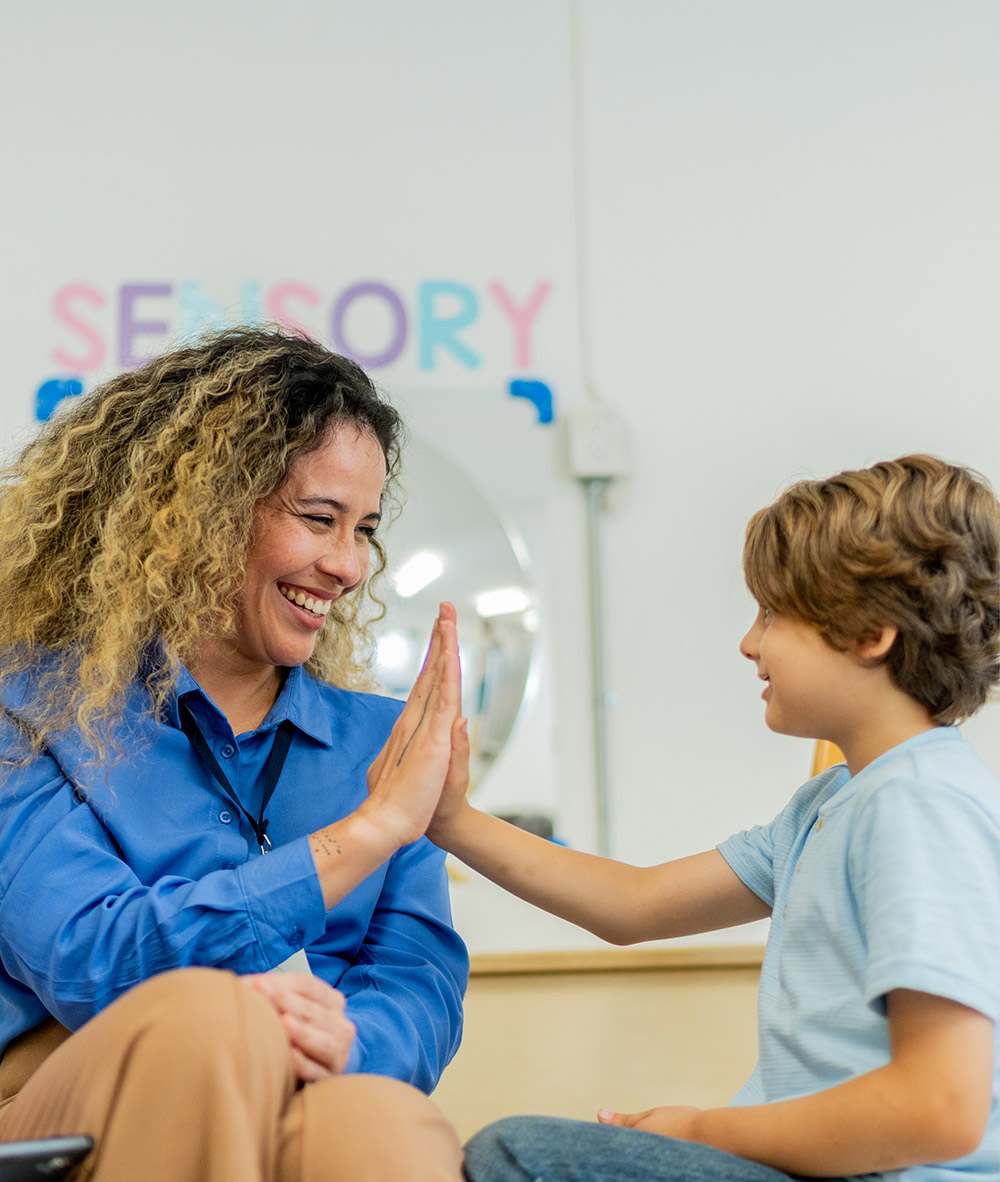 Smiling teacher giving a high five to a young boy in a sensory classroom.