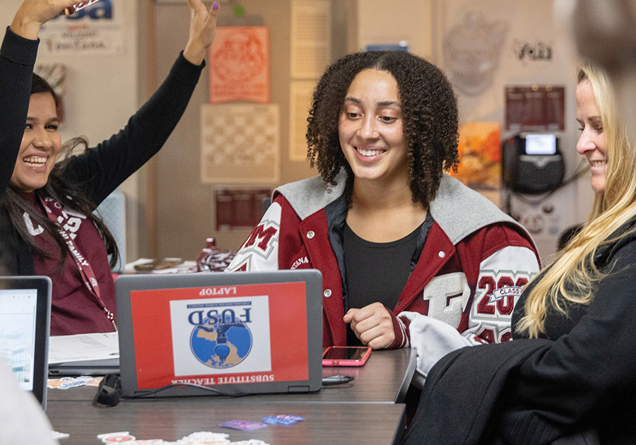 A high school student in a letterman jacket sitting at a laptop while celebrating with others.