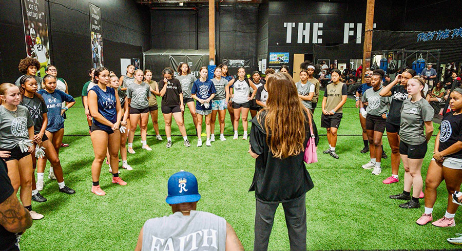 A coach addressing a large group of female athletes gathered on indoor turf.