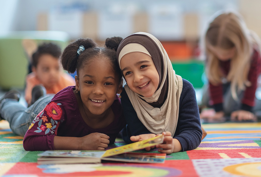 Two young girls lie on a colorful rug reading a book together in a classroom.