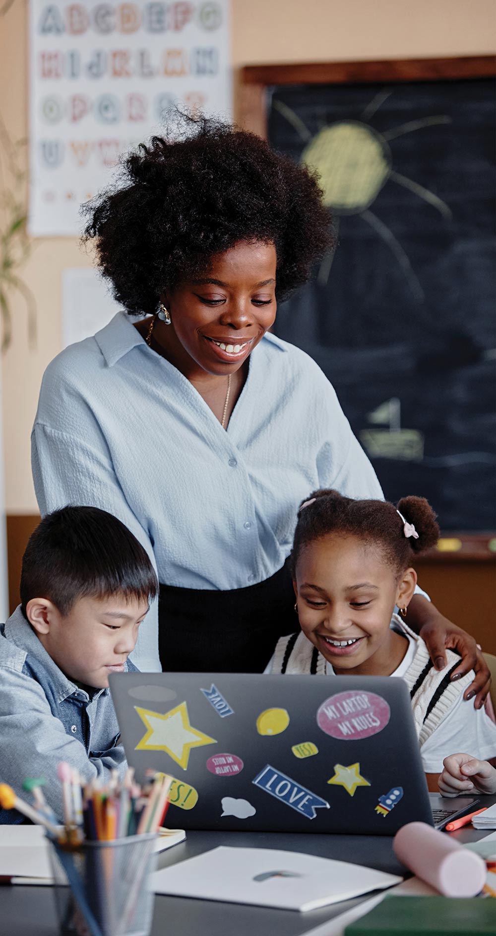Teacher smiling as she guides two young students working on a laptop in class.