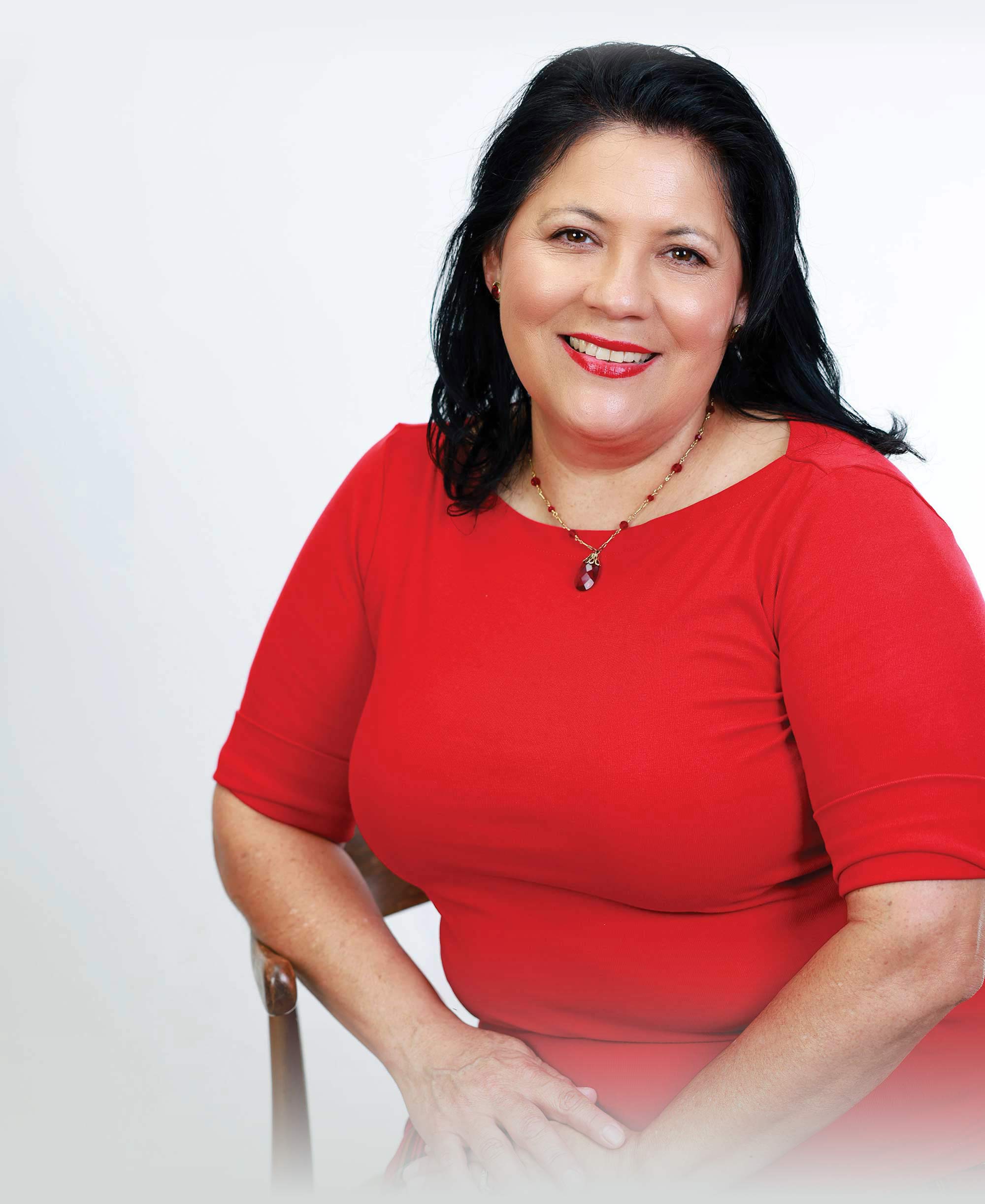 A portrait of Sonia Heredia smiling while seated in a red dress against a white backdrop.