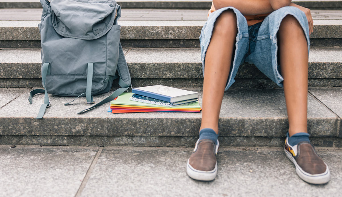 A student sitting on outdoor stone steps next to a grey backpack and a stack of colorful notebooks and folders.