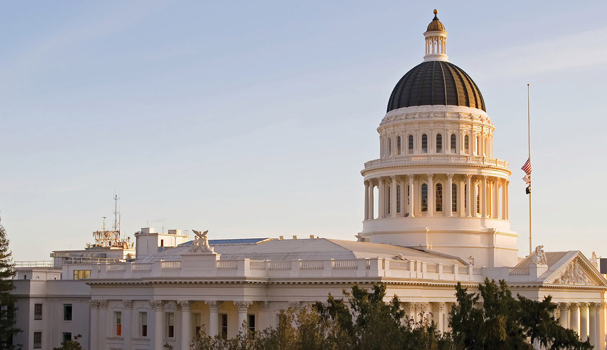 Exterior view of the California State Capitol building featuring its white facade, dark dome topped with a gold cupola, and trees in the foreground under a clear sky.