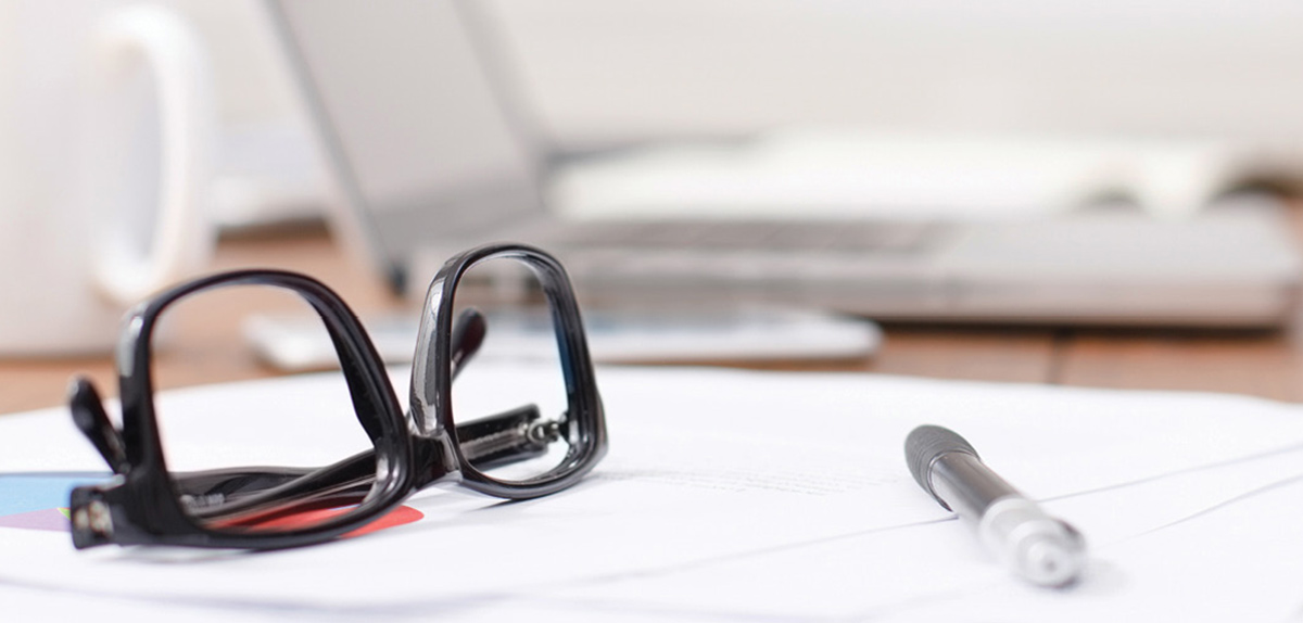 Close-up of black-framed eyeglasses and a pen resting on a printed document, with a blurred laptop and mug in the background on a wooden desk setup.