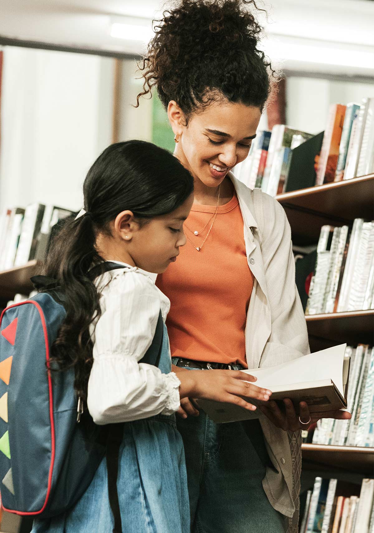 Woman and young girl with a backpack looking at an open book together in a library.